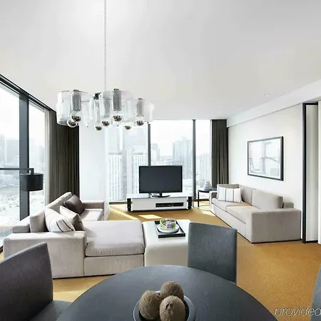 Crown Metropol Melbourne Hotel Room photo The photo shows a modern, stylish living space. The room features large windows that provide a panoramic view of the city skyline. There is a comfortable light-colored sofa arranged in the corner, complemented by a dark floor lamp. Across from the so